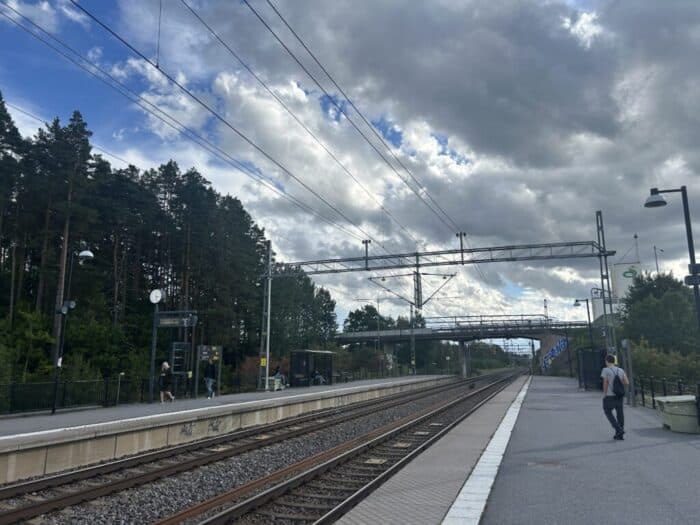 Empty train station platform with overhead electric lines, trees in the background, and cloudy sky, capturing a serene moment in urban transportation infrastructure.