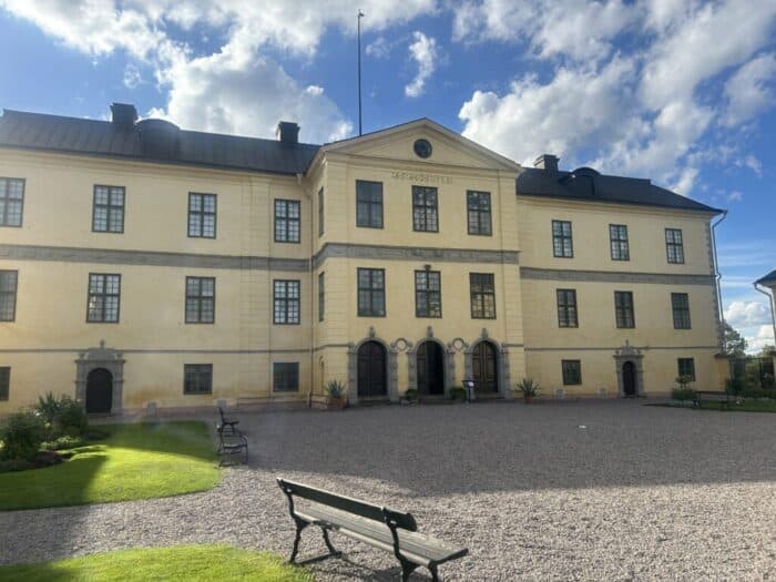 Historic yellow building with a manicured garden and benches, under a bright sky, showcasing classic architecture and well-kept outdoor space, perfect for moments of reflection.