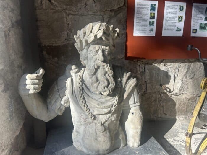 Ancient stone bust sculpture of a bearded man wearing a laurel wreath, displayed indoors with informational posters on the wall behind.